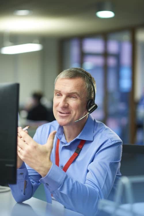 a call centre phone operative in his mid 40s chats on the phone at his desk . He is explaining something to the person on the phone in a friendly manner . behind him a defocussed office interior can be seen . This could be a call centre or an office worker chatting to a customer .