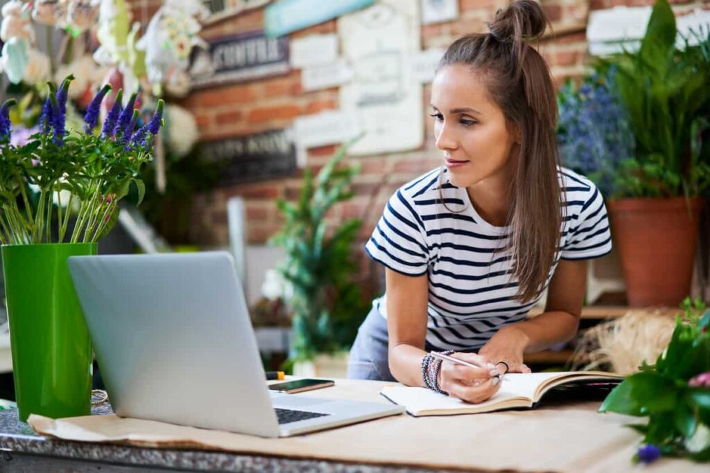 Young florist writing in notebook and looking at laptop while leaning on counter
